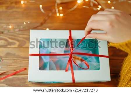 First-person view of woman's hand in knitted sweater opening Christmas surprise. Girl pulls ribbon on gift box. Wooden table with garland. New Year's holiday mood.
