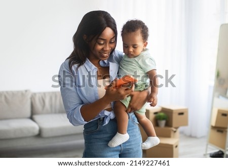 Portrait of smiling young African American woman showing educational app on cell phone to cute small son, lady holding baby on hands, female using digital gadget, watching videos together