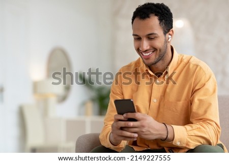 Smiling Arabic Male Listening To Music On Mobile Phone Wearing Wireless Headphones Sitting On Couch Relaxing At Home. Male Enjoying Favorite Playlist Using Musical Application, Free Copy Space Royalty-Free Stock Photo #2149227555
