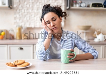 Sleepy young woman sitting at dining table in kitchen with closed eyes, holding cup while having breakfast. Early wake up, insomnia, lack of sleep Royalty-Free Stock Photo #2149227681