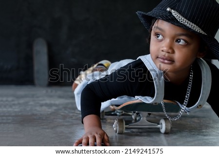 Close up little African kid boy wear hat, necklace chain and jeans bib practice playing and lying on skateboard pushing cement floor on black background. Cool child playing extreme sport on free time