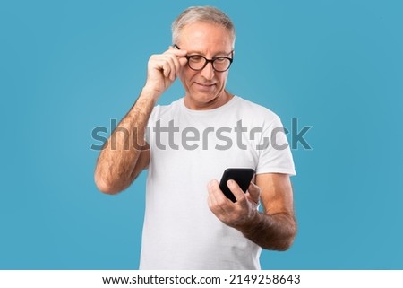 People And Technology. Portrait of happy smiling mature man using smartphone and touching putting on glasses standing isolated over blue studio wall. Adult male chatting online, browsing social media Royalty-Free Stock Photo #2149258643