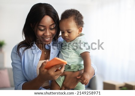 Cheerful black mother holding kid on hands showing smart phone to little son, using entertainment or educational application having fun at home. Concept of children development, parental control