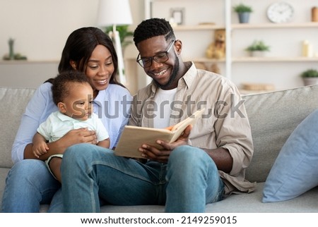 Leisure With Child. Caring Young Parents Reading Book To Their Adorable Infant Son Relaxing Together Sitting On Couch In Living Room, Happy Dad And Mum Bonding With Toddler Baby Kid At Home