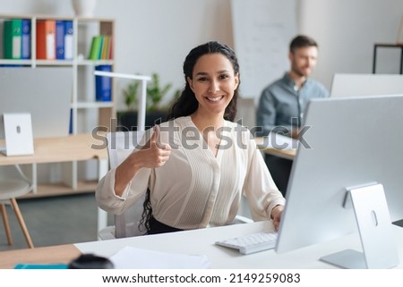 Cheerful young female employee showing thumb up gesture while working on computer in modern office. Happy millennial secretary expressing her agreement or approval, positive evaluation Royalty-Free Stock Photo #2149259083
