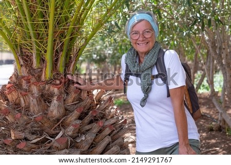Portrait of beautiful active elderly woman with backpack on outdoor hike resting in the shade of a palm tree. Elderly sporty female enjoying healthy lifestyle in nature