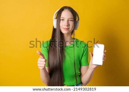 Portrait young teen girl posing isolated over yellow background wall listening to music with wireless headphones using mobile phone in bright green t-shirt. Mockup screen