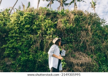 Side view of young female traveler wearing casual clothes standing near hill and taking picture on photo camera while spending time in tropical country