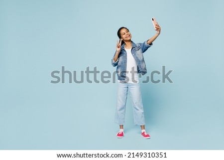 Full body little kid teen girl of African American ethnicity 12-13 years old in denim jacket do selfie shot on mobile cell phone show v-sign isolated on plain light blue background. Childhood concept