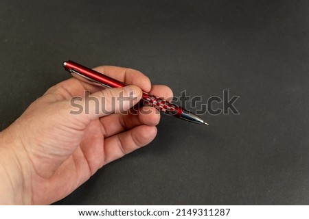 A close-up of a man's hand holding a red pen against a black background. A mature businessman with a metal ballpoint pen in his left hand. Royalty-Free Stock Photo #2149311287
