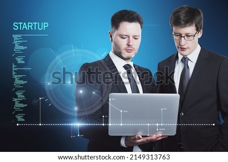 Attractive young european businessmen using laptop with futuristic blue HUD interface and glowing programming code on blurry blue background. Digital start up, teamwork, future and technology concept