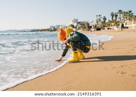 Boy in yellow rubber boots playing with water and sand at the beach. School kid touching water at autumn winter sea. Child having fun with waves at the shore. Spring Holiday vacation concept.