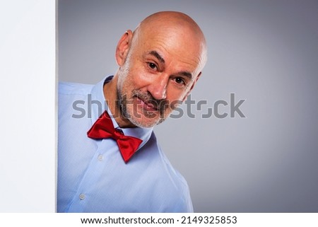Studio shot of funny faced middle aged man looking out of white board while standing at isolated grey background. Copy space. 