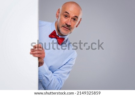 Studio shot of funny faced middle aged man looking out of white board while standing at isolated grey background. Copy space.  Royalty-Free Stock Photo #2149325859