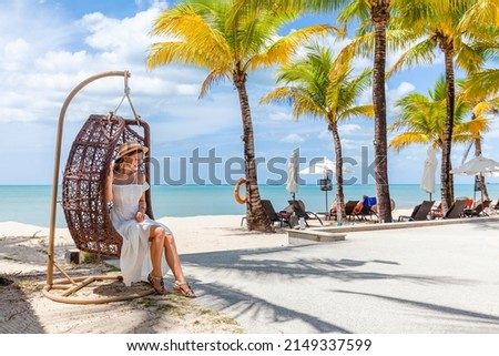 Young attractive woman in straw hat rest in rattan wicker cocoon swing chair by beach in luxury beachfront hotel resort at sunny day, enjoy holiday vacation. Sea, blue sky, palm trees on background. Royalty-Free Stock Photo #2149337599