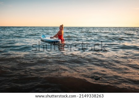 Pretty adult plump woman in a red swimsuit swimming in the sea with a sup board. Copy space. The concept of active recreation and sports.