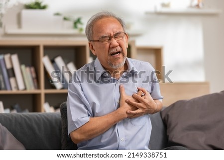Elderly Asian male with from heart attack sitting at the couch in the living room. senior clutching his left chest from acute pain. Heart attack symptom, he has difficulty breathing. Royalty-Free Stock Photo #2149338751