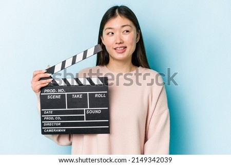 Young asian woman holding clapperboard isolated on blue background looks aside smiling, cheerful and pleasant.