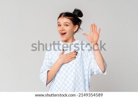 My name is. Smiling teen girl introduce herself, raising hand up and say hello. Little girl telling truth, swearing she is honest, making promise with palm on heart, light grey background