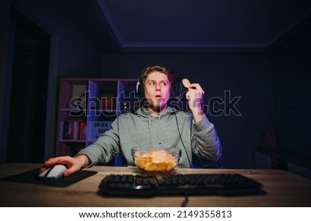 Positive young male gamer in casual clothes and headset playing online games on the computer and eating chips from a plate with a happy face.