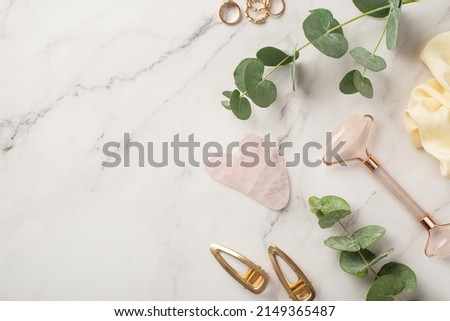 Skincare beauty procedure concept. Top view photo of gold barrettes rose quartz roller gua sha massager yellow scrunchy gold rings and eucalyptus on white marble background with copyspace