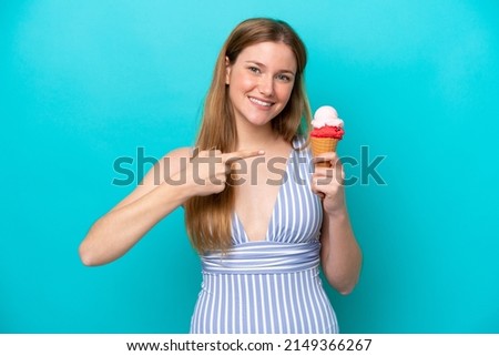 Young caucasian woman in swimsuit eating ice cream isolated on blue background and pointing it