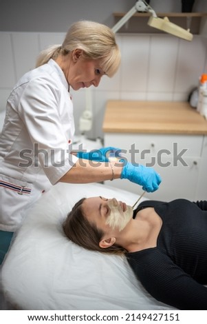 Doctor cosmetologist or dermatologist making face mask in cosmetology salon. Professional Beautician applying face mask on caucasian woman face lying on bed in bathrobe. High quality photo