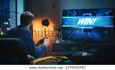 Young Man Spending Time at Home, Sitting on a Couch in Stylish Loft Apartment and Playing Arcade Car Video Games on Console. Male Using Controller to Play Street Racing Drift Simulator.