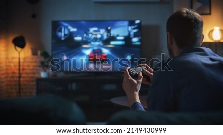 Young Man Spending Time at Home, Sitting on a Couch in Stylish Loft Apartment and Playing Arcade Car Video Games on Console. Male Using Controller to Play Street Racing Drift Simulator. Royalty-Free Stock Photo #2149430999