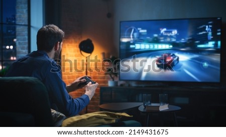 Young Man Spending Time at Home, Sitting on a Couch in Stylish Loft Apartment and Playing Arcade Car Video Games on Console. Male Using Controller to Play Street Racing Drift Simulator. Royalty-Free Stock Photo #2149431675