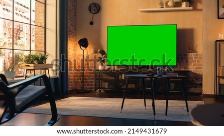 Stylish Loft Apartment Interior with TV Set with Green Screen Mock Up Display Standing on Television Stand. Empty Living Room at Home with Chroma Key Placeholder on Monitor. Zoom In Sunset Shot. Royalty-Free Stock Photo #2149431679