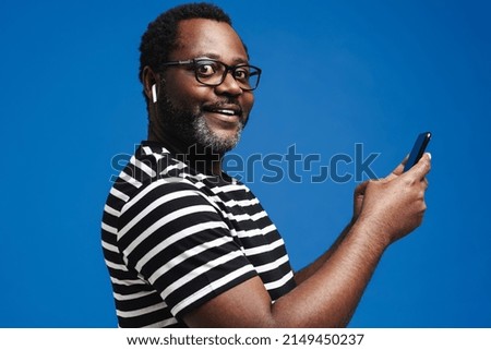 Black man in earphones listening music while using mobile phone isolated over blue background