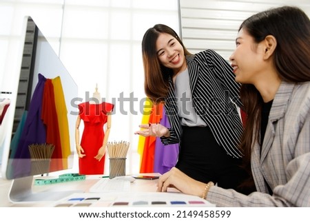 Two Asian young happy success female stylish fashion designer wears casual suit talk discuss creative design information and point at computer monitor together in front of clothes rack and mannequin.