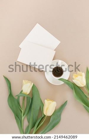 Blank paper sheet cards, white cup of coffee, pastel yellow tulips on beige background. Mock up. Copy space. Minimal business brand template.