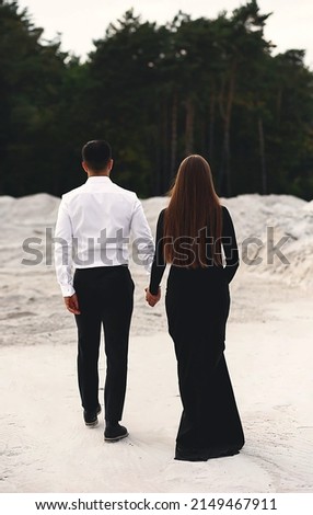 Rear view young happy lovely couple, man and woman in black dress hold hands walking stroll together near forest and looking into the distance. Nature background. Love concept