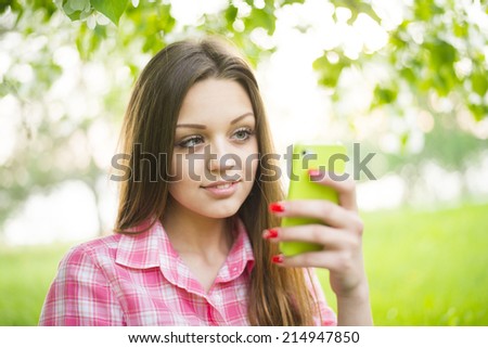 Caucasian beautiful girl looking at camera against fresh spring green grass and leaves of summer trees background Close up portrait of Young adult woman using green mobile cell phone in park 