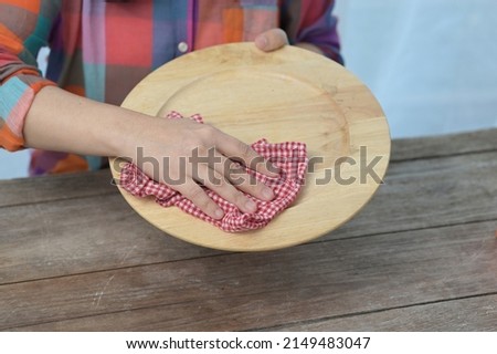 The woman's right hand holds a red (plaid) dishcloth, wipes a large plate made of wood before placing it in a dish rack. A housewife in brightly colored clothes is cleaning the dishes.