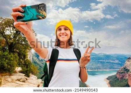 Portrait of young cheerful woman backpacker with short brunette hair and yellow beanie, taking selfie with smartphone doing victory sign with hand over the top of the mountain lake. Adventure, travel.