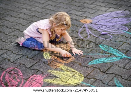 Little preschool girl painting with colorful chalks flowers on ground on backyard. Positive happy toddler child drawing and creating pictures on asphalt. Creative outdoors children activity in summer.