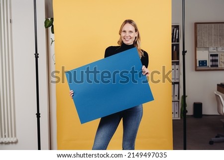 young woman stands in front of yellow background and shows a blank blue sign for advertising