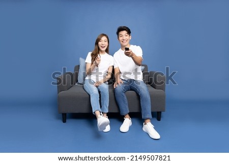 Young Asian couple holding remote tv and watching television on a sofa isolated on blue background Royalty-Free Stock Photo #2149507821