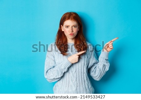 Winter holidays and people concept. Complicated redhead girl staring indecisive, pointing fingers right at logo and frowning, standing perplexed against blue background