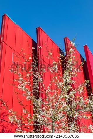 The wall of the building with triangular projections. Modern architecture in red color. Abstract geometric pattern building with spring tree. Street photo, selective focus, nobody.