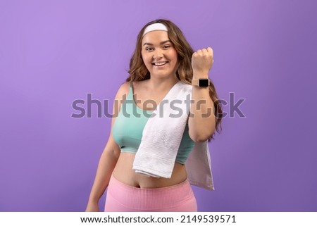 Glad young european plus size female in sportswear with towel show fit tracker after workout, isolated on purple background. Excellent result, fitness and training, cardio exercises and motivation Royalty-Free Stock Photo #2149539571
