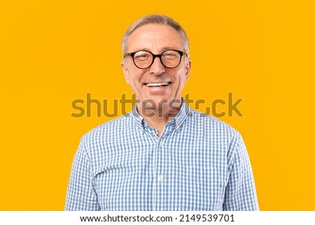 Portrait of senior male smiling wearing glasses and casual checkered shirt posing standing isolated over yellow orange studio background. Happy excited adult person looking at camera, free copy space
