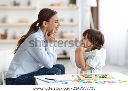 Speech therapy concept. Professional woman specialist training with cute little boy, showing him oral exercise to warm up mouth muscles, side view Royalty-Free Stock Photo #2149539733