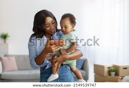 Modern Technology. Happy smiling African American mum holding little son on hands standing in living room, using cell phone watching cartoons together, mom showing kid educational online program