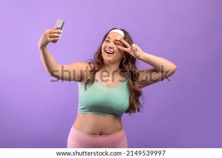 Cheerful millennial european plus size lady in sportswear taking selfie on phone and shooting livestream, isolated on purple background, studio. Photo, app for fitness blog and website, modern tech