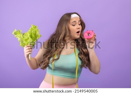 Hungry young funny european plus size woman in sportswear with salad in her hand eats donut, isolated on purple background, empty space. Health care, weight loss and diet, sweet tooth and motivation