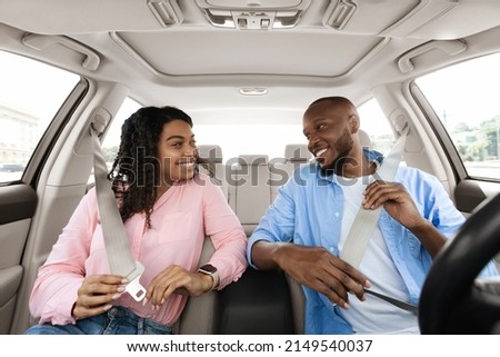 Smiling African Amerfican couple fastening seat belts after get in brand new car, happy black lovers having weekend trip together, ready for vacation, driving modern auto, windshield view. Safety Fist Royalty-Free Stock Photo #2149540037